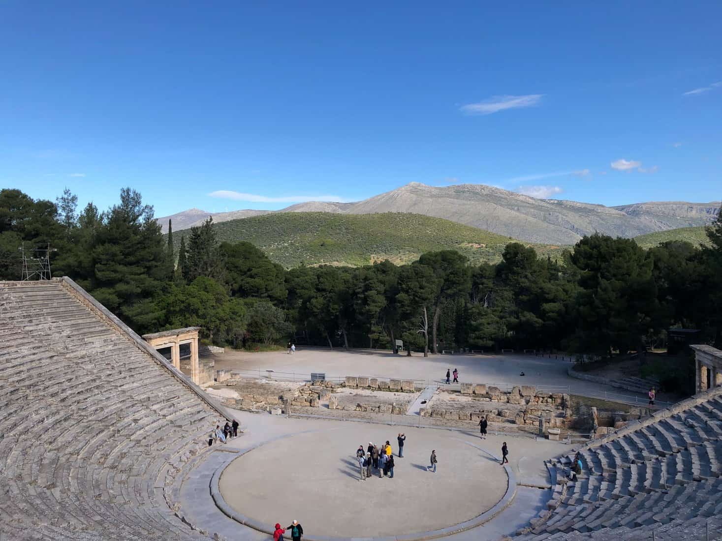 A distant blue sky, low-sloped, grey and green mountains, all preceding the ancient Greek amphitheatre of Epidaurus (Peloponnese, Greece, 2019).
